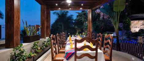 An outdoor dining area with wooden chairs and tables, surrounded by plants and a view of the night sky.