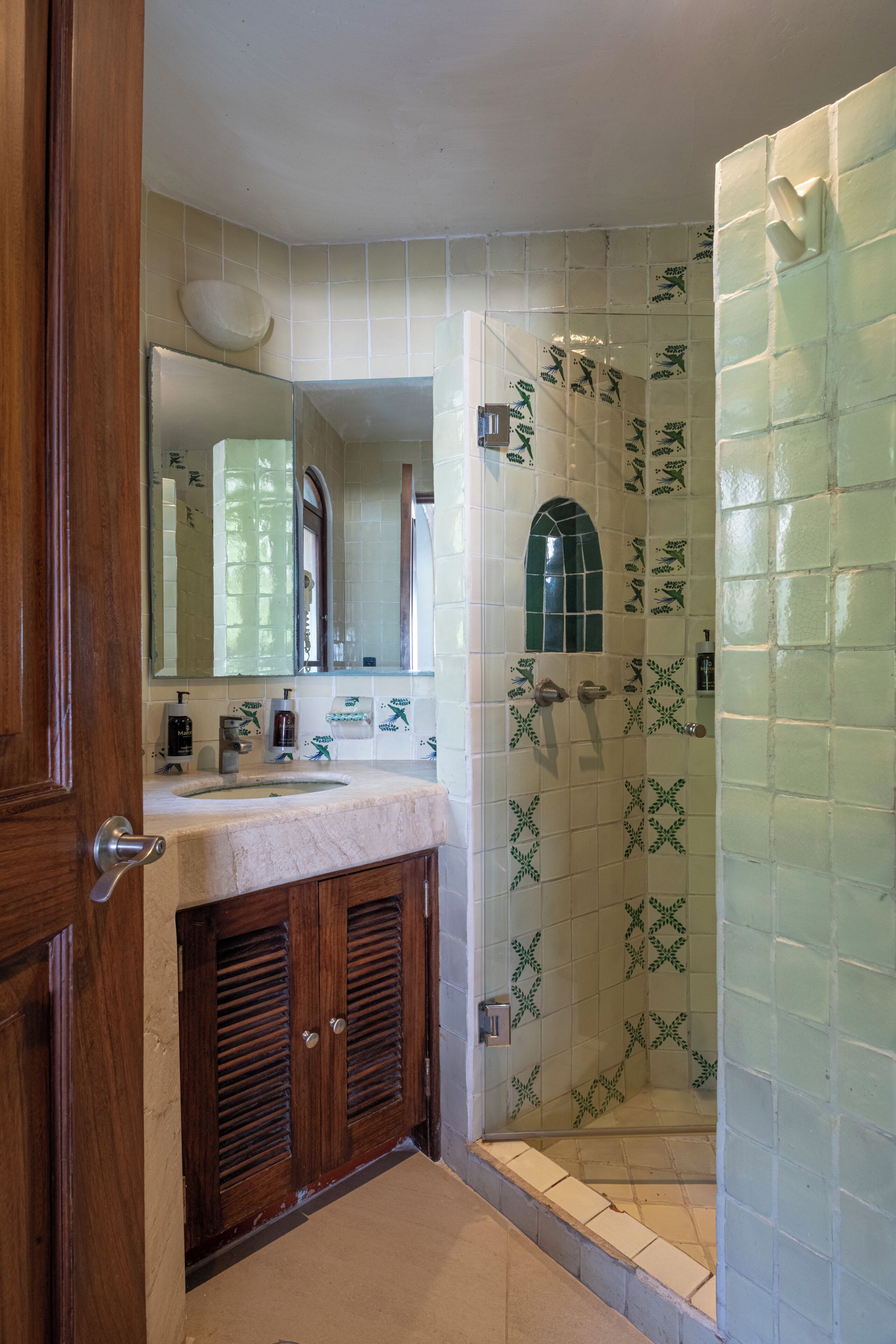 Une salle de bain avec un meuble-lavabo en bois, un évier, un miroir et une douche dont les murs sont recouverts de carreaux à motifs floraux verts.