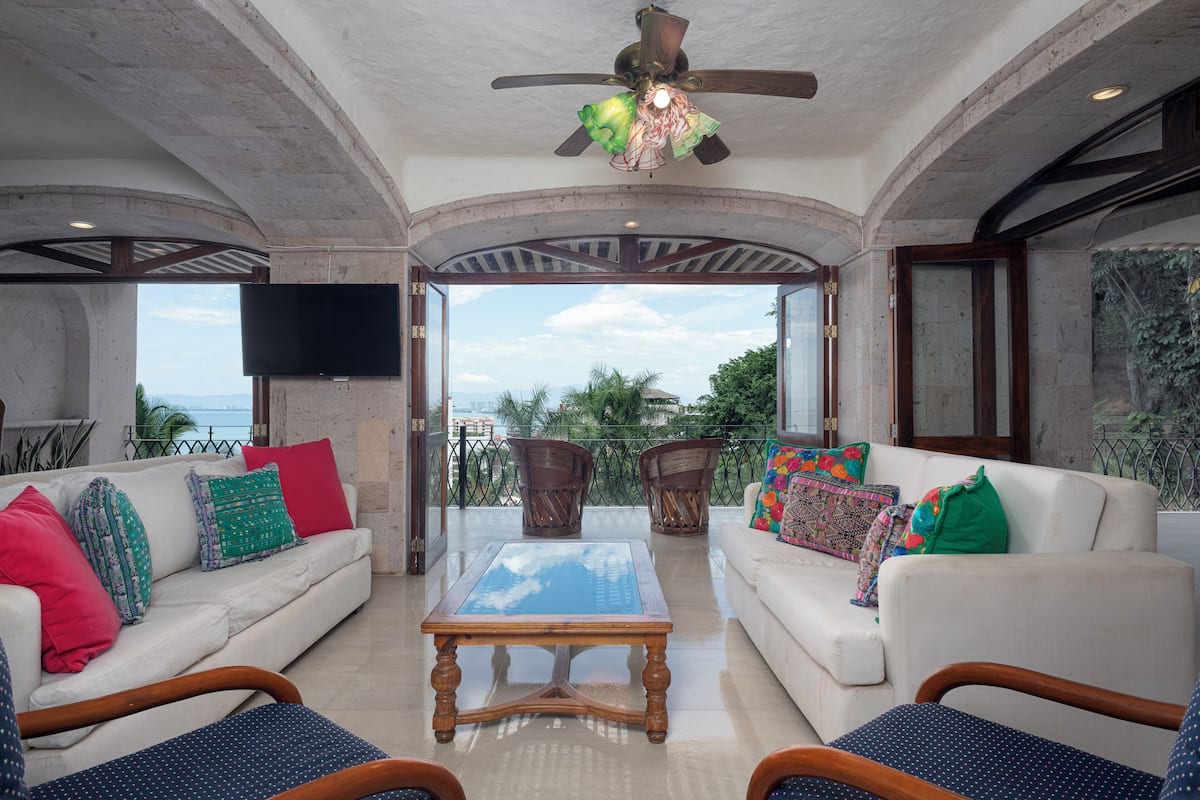 A spacious living area with a ceiling fan, two sofas with colorful cushions, a wooden coffee table, and a view of the outdoors.