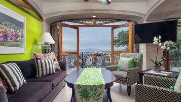 Une pièce comprenant un canapé, des fauteuils, une table recouverte d’une nappe verte, un téléviseur et un balcon avec vue.