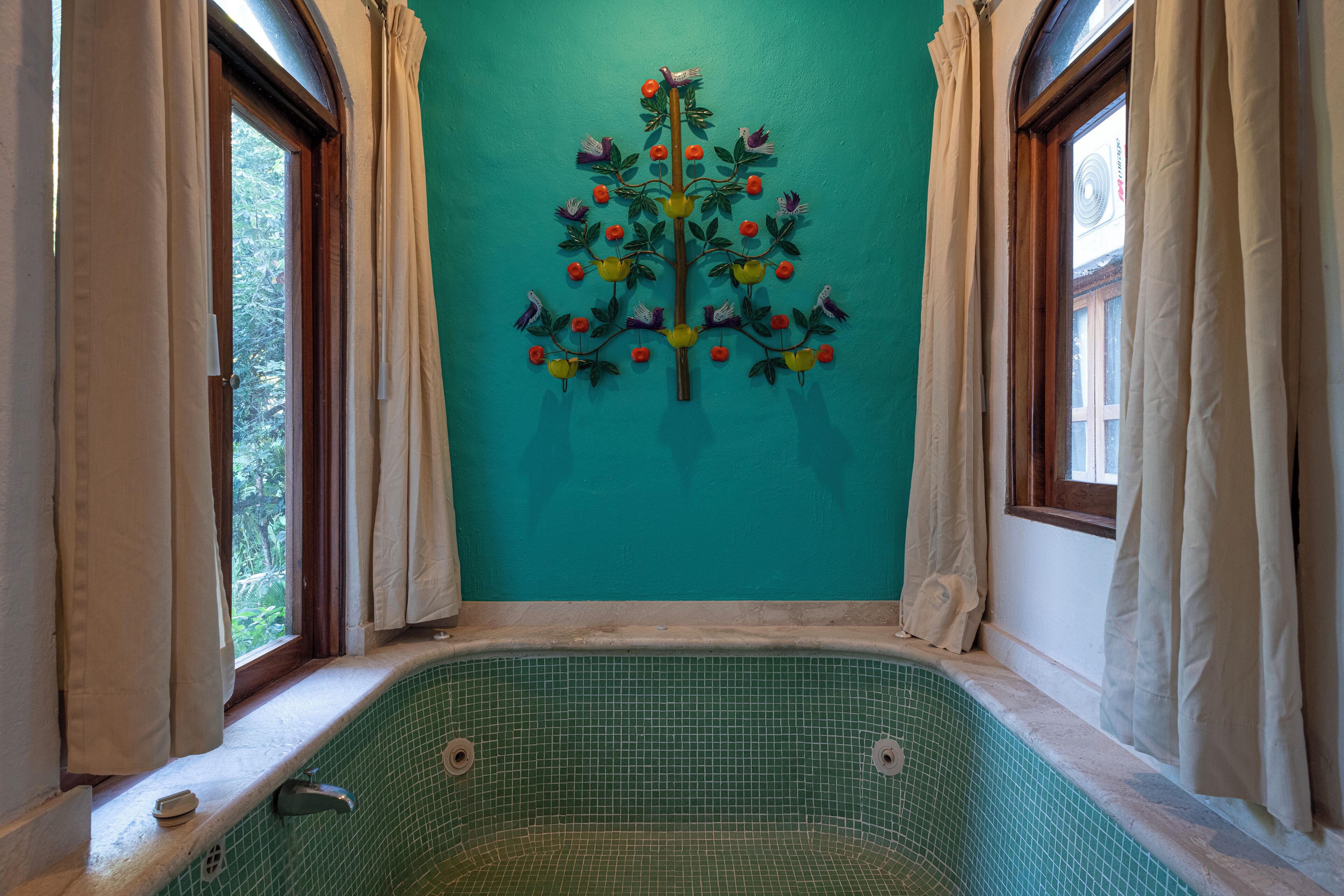 Una habitación con una bañera de azulejos verdes, un adorno con forma de árbol en la pared y ventanas con cortinas.