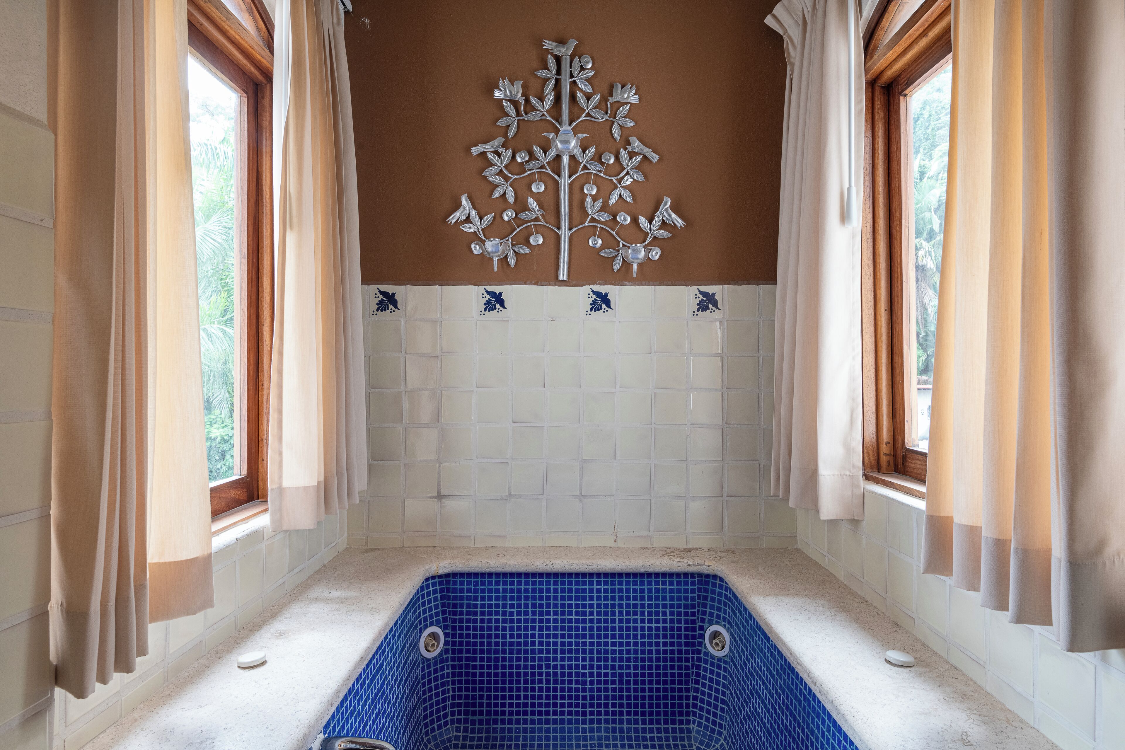 Une salle de bain avec une baignoire carrelée de carreaux bleus, des rideaux beiges et un élément décoratif mural.