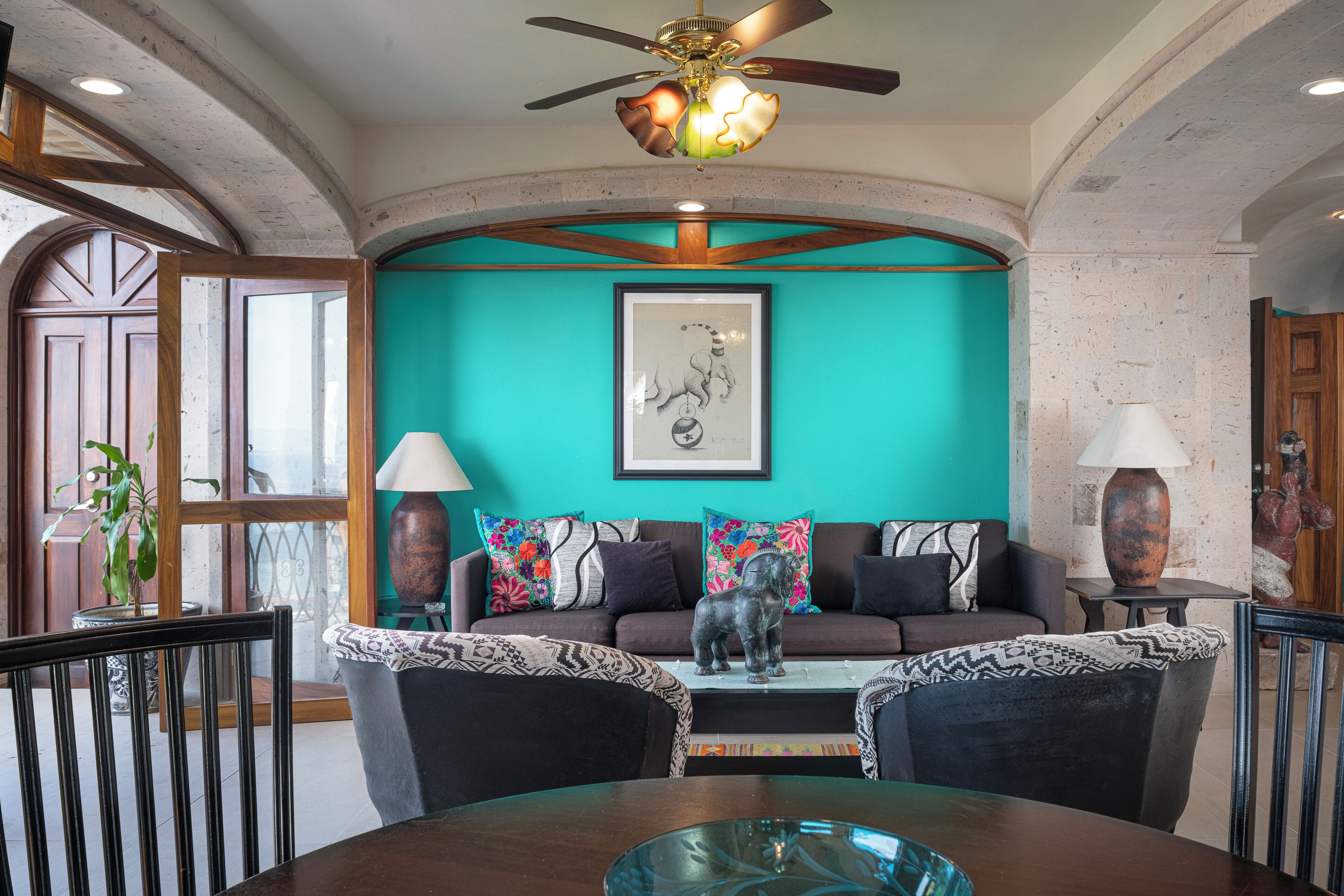 A living room with a teal wall, a ceiling fan, a framed artwork, and a sofa with patterned cushions.