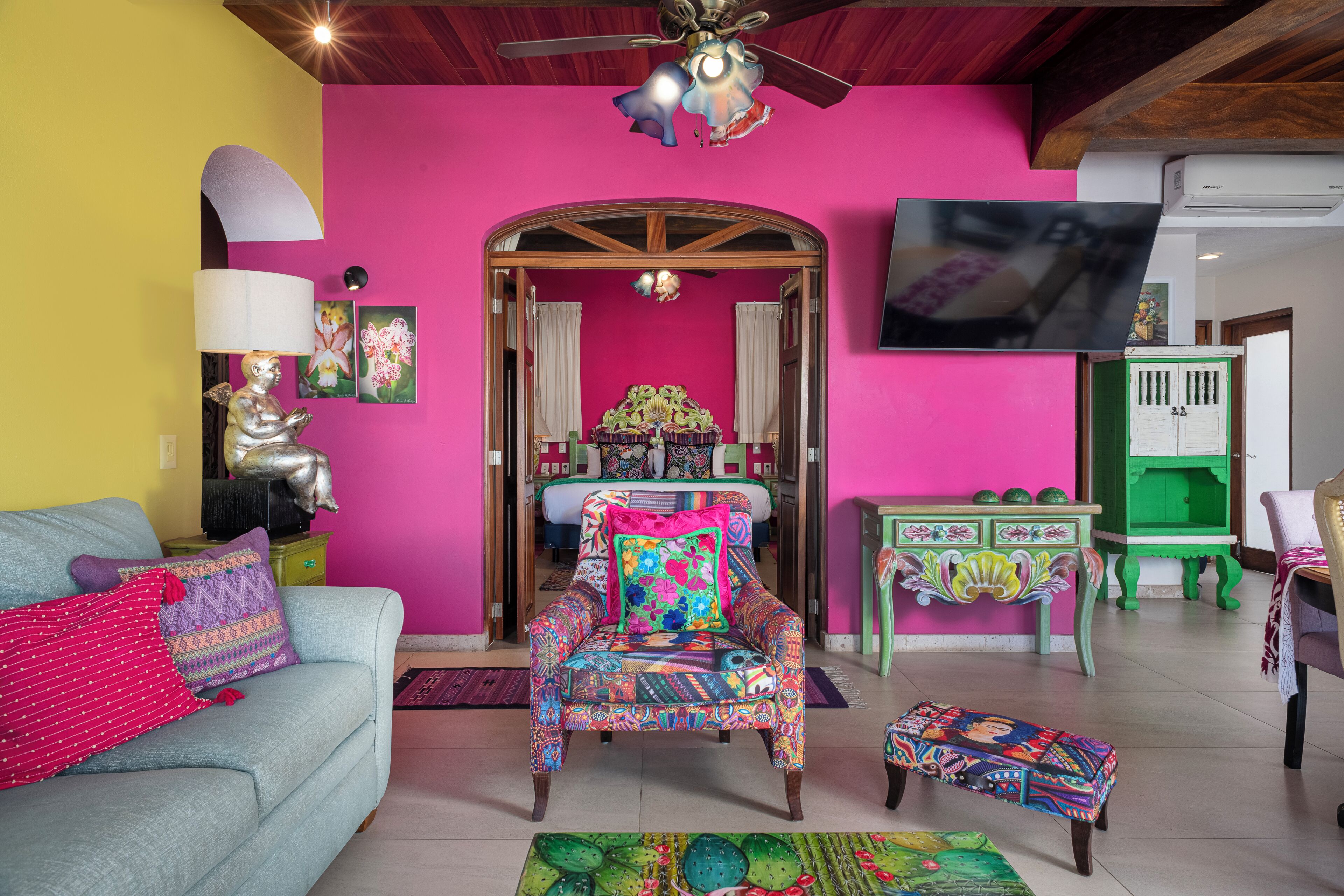 A room with a sofa, a colorful armchair, a small table, and a television mounted on the wall.