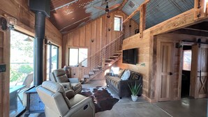 A spacious living area with a high ceiling, wooden walls, a large window, a sofa, a chair, a TV, and a staircase.