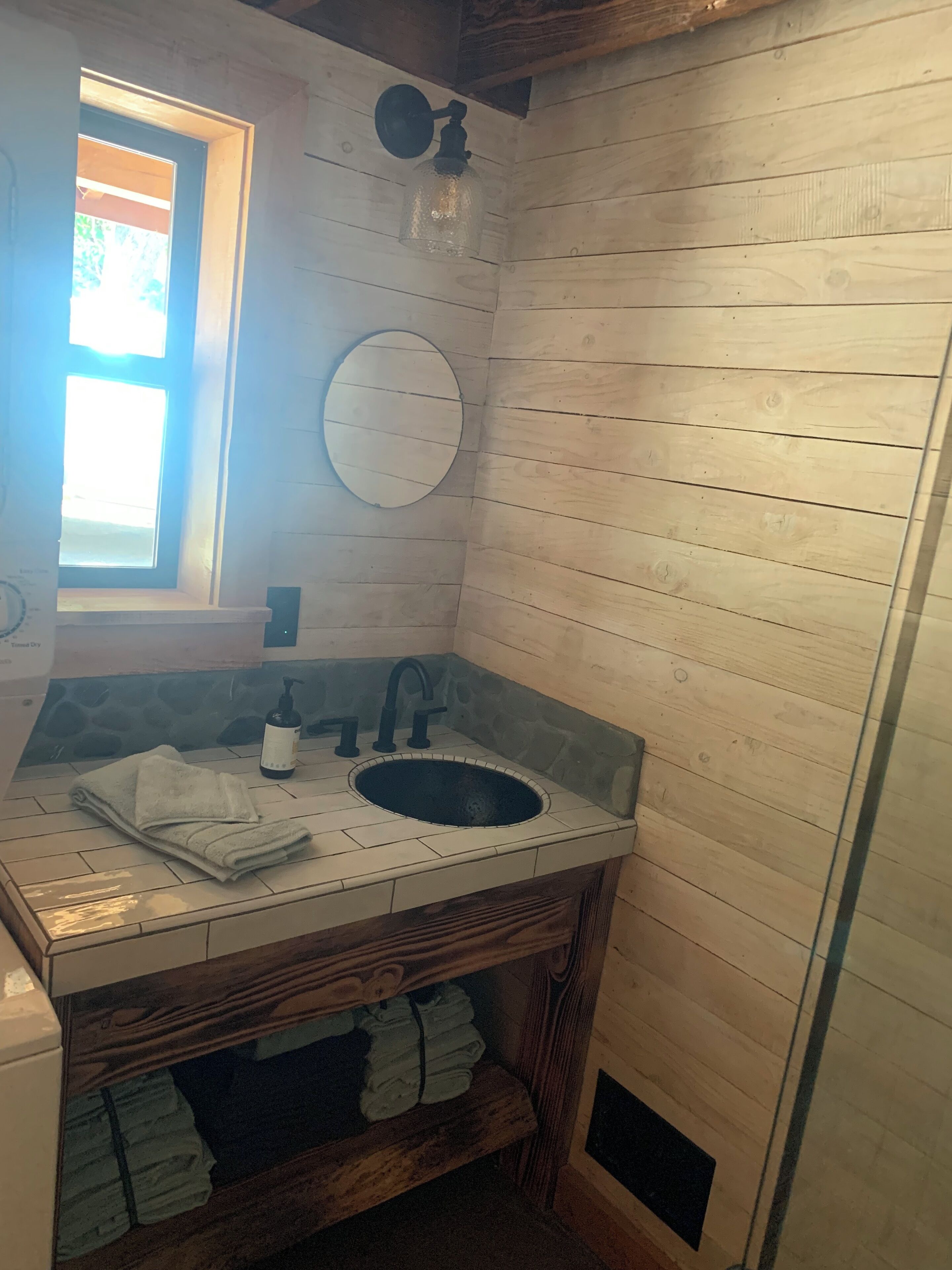 Un baño pequeño con pared de madera, un espejo redondo, un lavabo con grifo y una ventana.