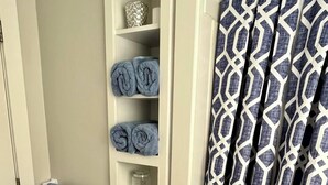 Une salle de bain avec un porte-serviettes sur lequel sont posées deux serviettes, une étagère avec des serviettes pliées, un bocal en verre, un miroir rond et un rideau de douche à motifs.