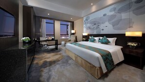 Una habitación de hotel moderna con una cama grande, un escritorio y un televisor. Hay ventanas con cortinas, un cuadro en la pared y suelo alfombrado.
