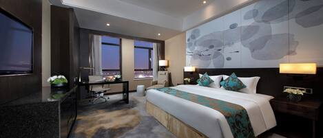 A modern hotel room with a large bed, a desk, and a TV. There are windows with curtains, a painting on the wall, and a carpeted floor.