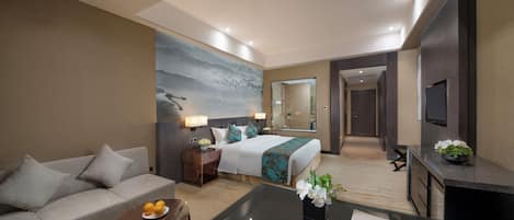 Une chambre d’hôtel moderne avec un canapé, un lit agrémenté d’oreillers décoratifs, une petite table avec des fruits, une table basse en verre et un téléviseur fixé au mur.