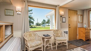 A room with two wicker chairs, a small table, a wooden dresser, and a window with a view of a green landscape. - Cottage On the Bay - Queen, Jetted tub, Full Kitchen, water view (Sequim)