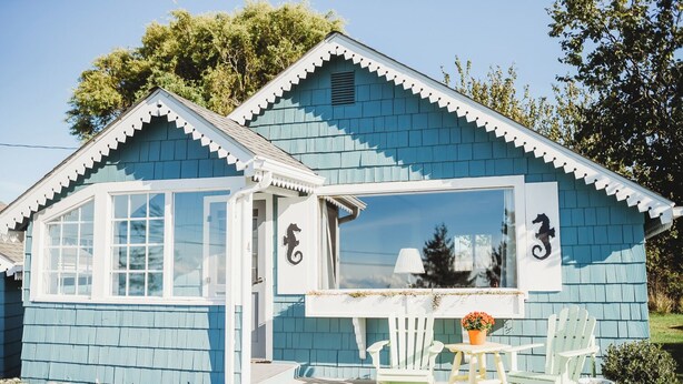 A small, single-story house with a blue exterior, white trim, and a large window. There is a white door with a seahorse decal, a small porch with a table and two chairs, and a stone wall in front. - Cottage On the Bay - Queen, Jetted tub, Full Kitchen, water view (Sequim)