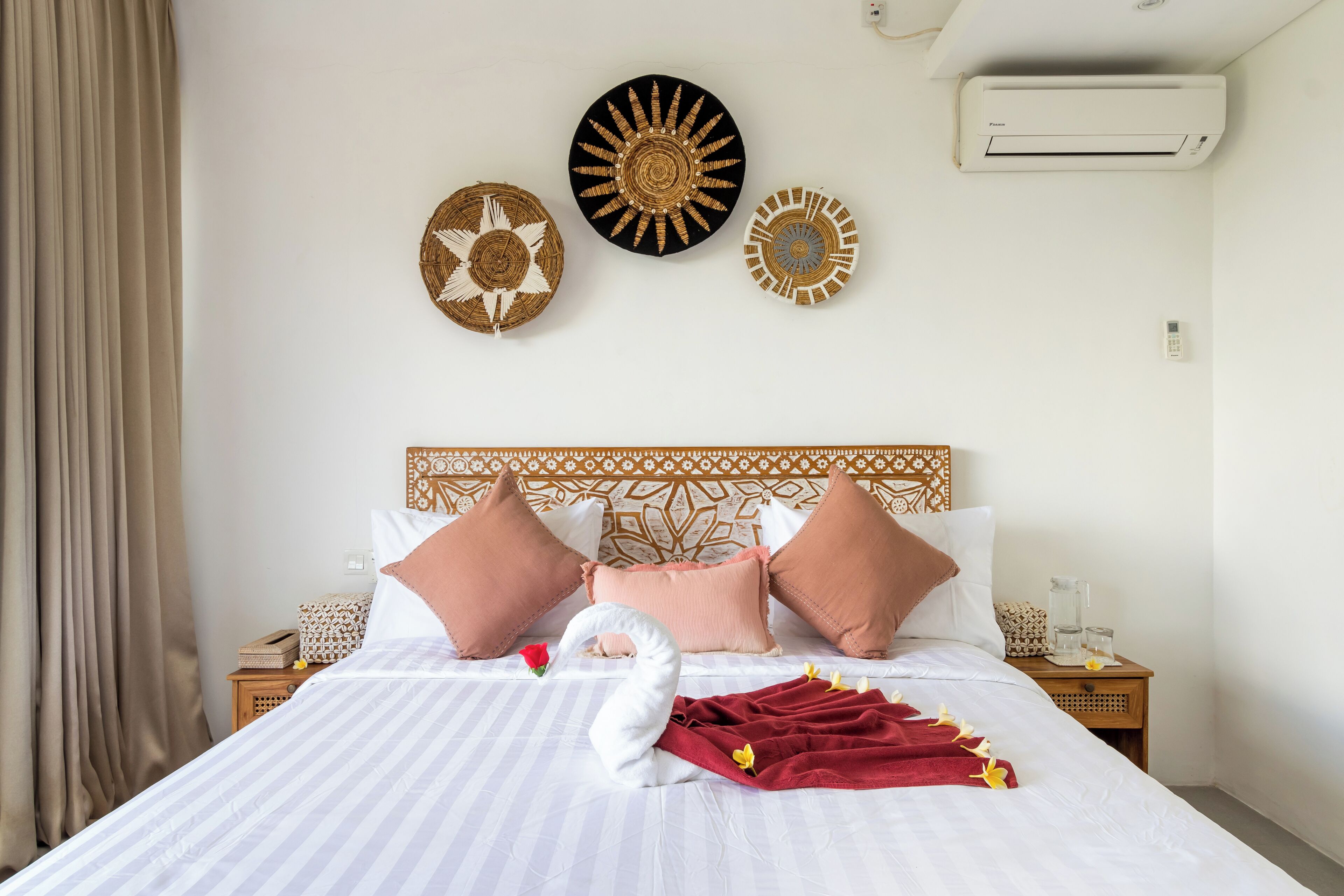 Un lit bien fait, recouvert d’une couverture blanche et rouge, agrémenté d’une serviette décorative et d’une rose rouge. Trois éléments décoratifs ornent le mur au-dessus du lit, ainsi qu’une tête de lit en bois et une table de chevet sur laquelle se trouvent un verre et un livre.