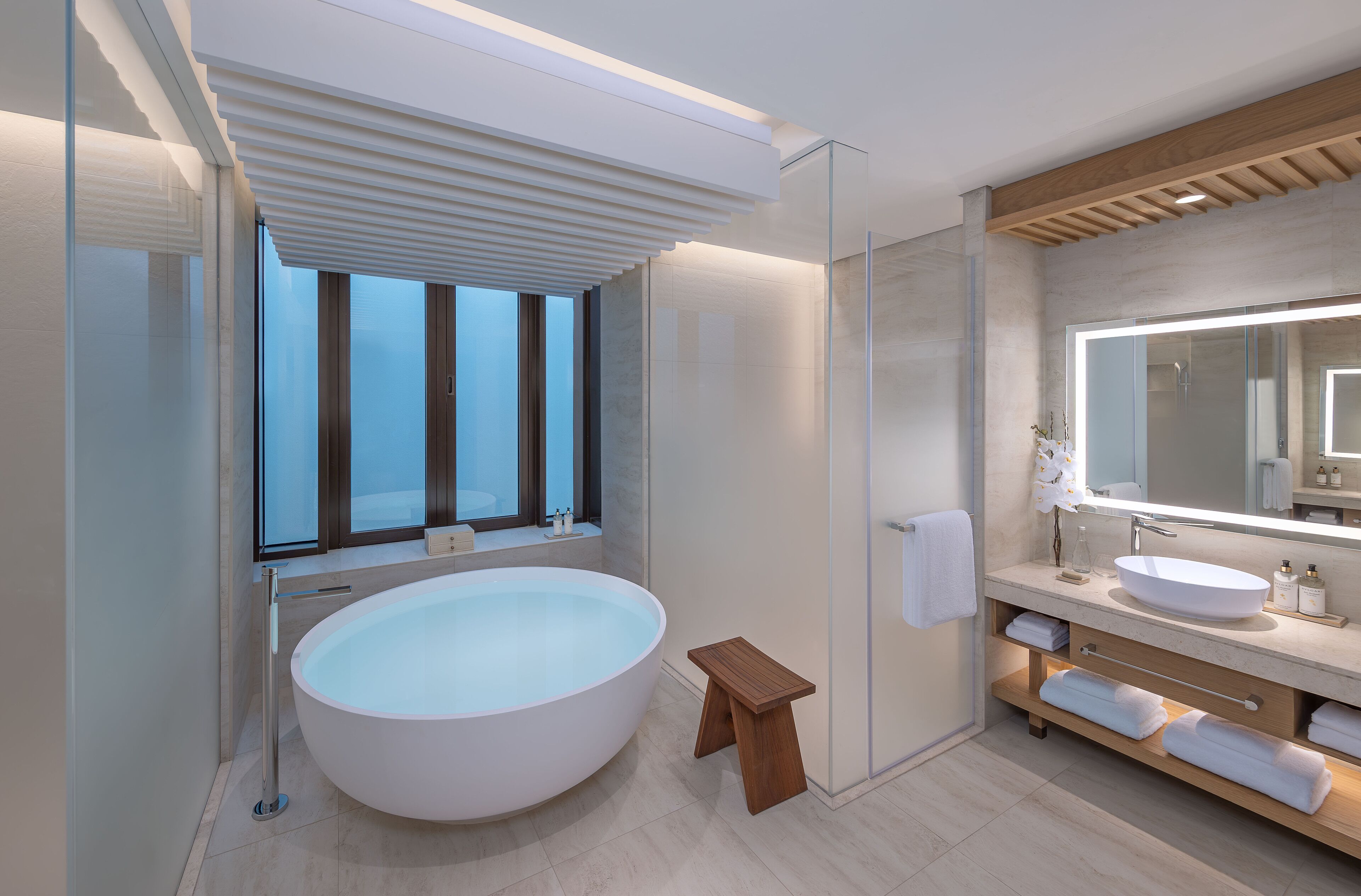 Un bagno moderno con una grande vasca da bagno ovale, un mobile bagno in legno, uno specchio grande e una finestra con persiane verticali.