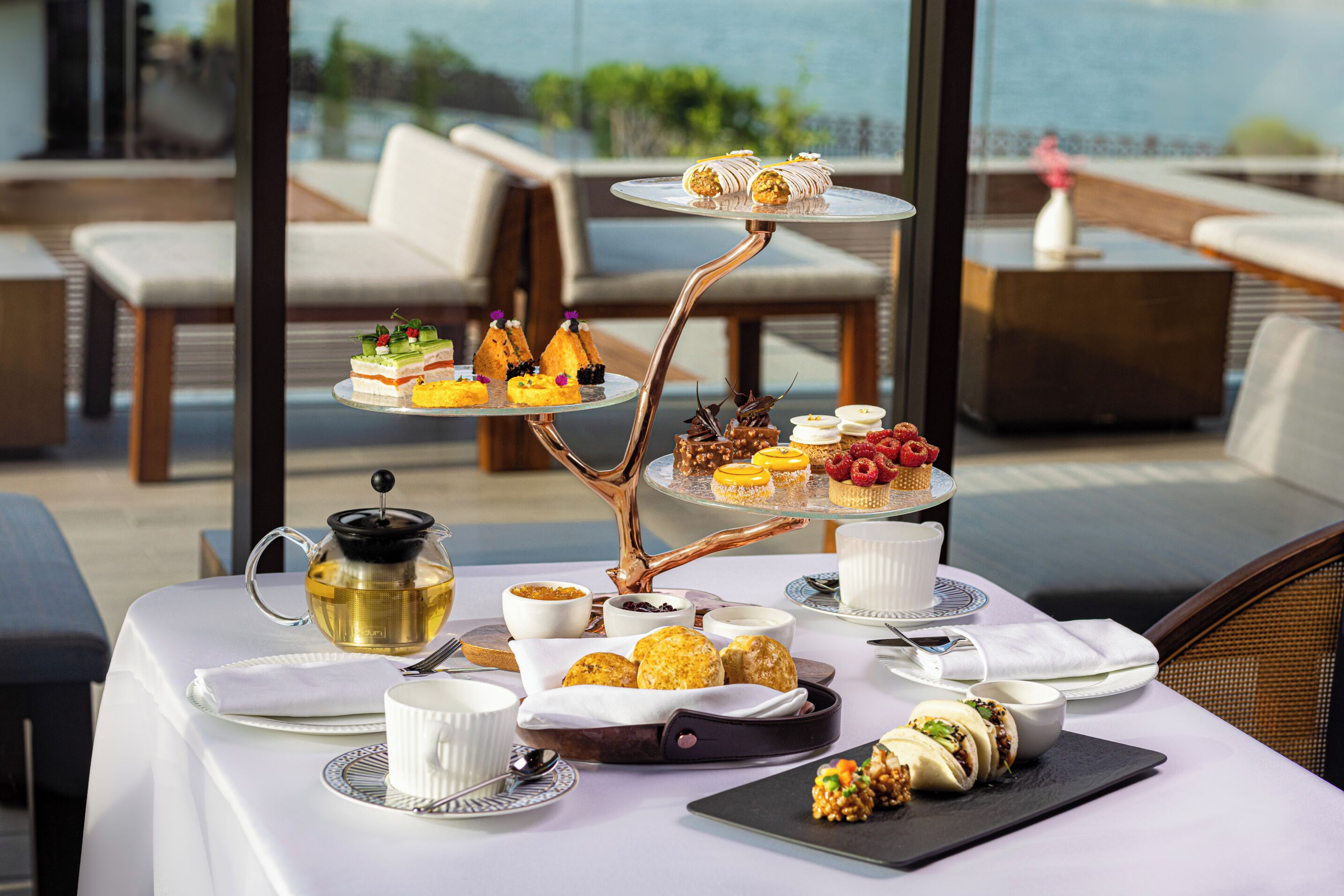 Une table dressée avec une variété de pâtisseries et de desserts, une théière et des tasses, sur une terrasse offrant une vue panoramique.