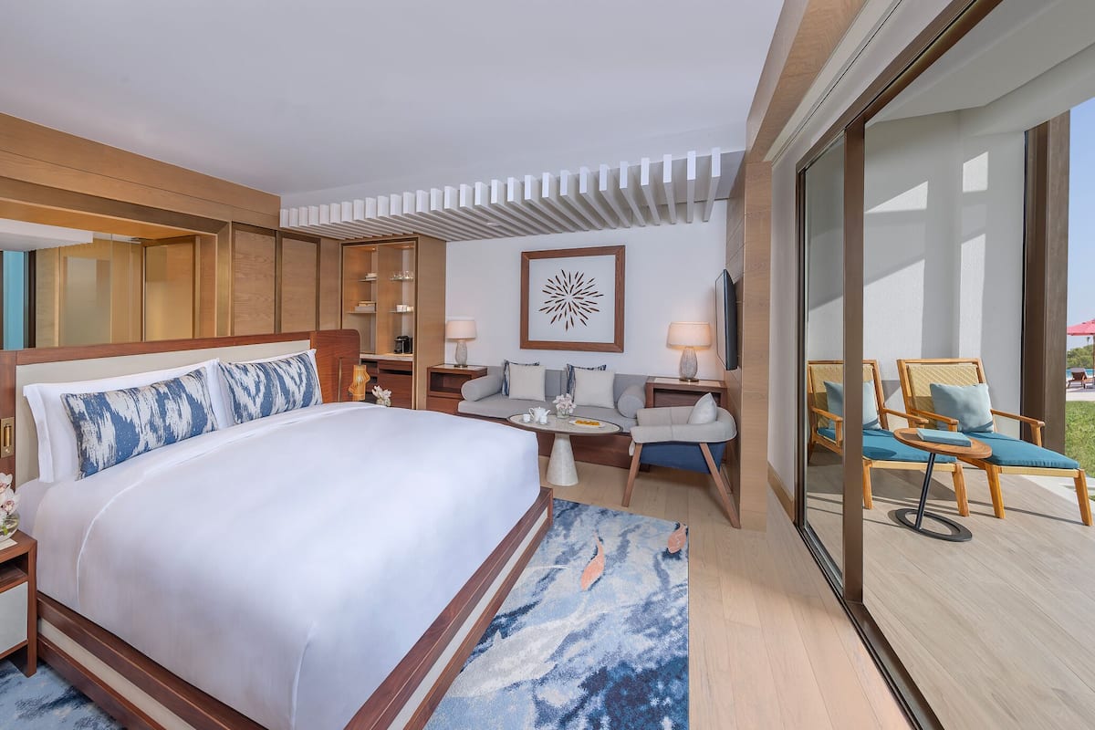 Una habitación de hotel moderna con una cama grande, una zona de estar con mesa y un balcón con mobiliario exterior.