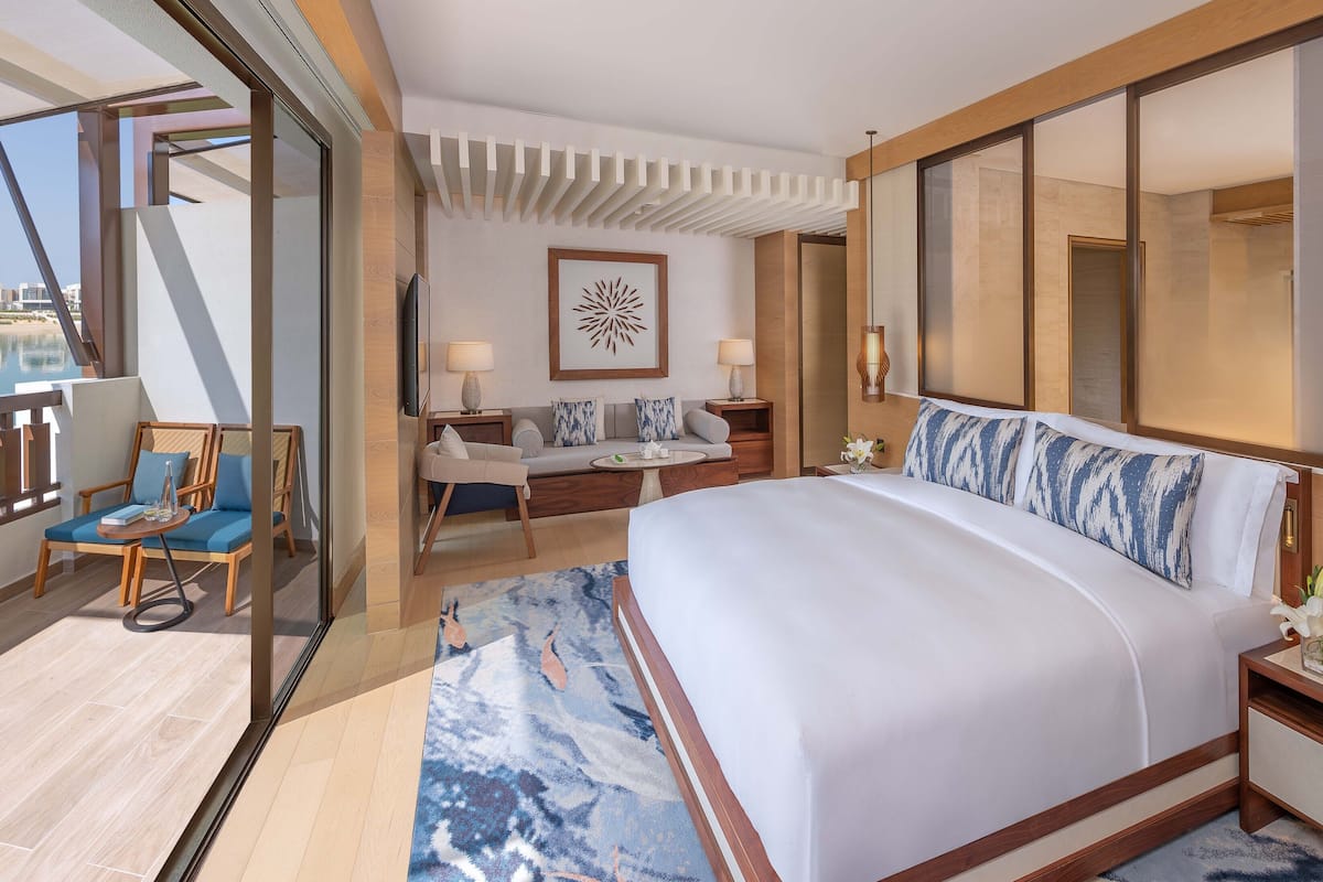 Una habitación de hotel moderna con una cama grande, un balcón con vistas, una zona de estar con mesa y un espejo amplio.