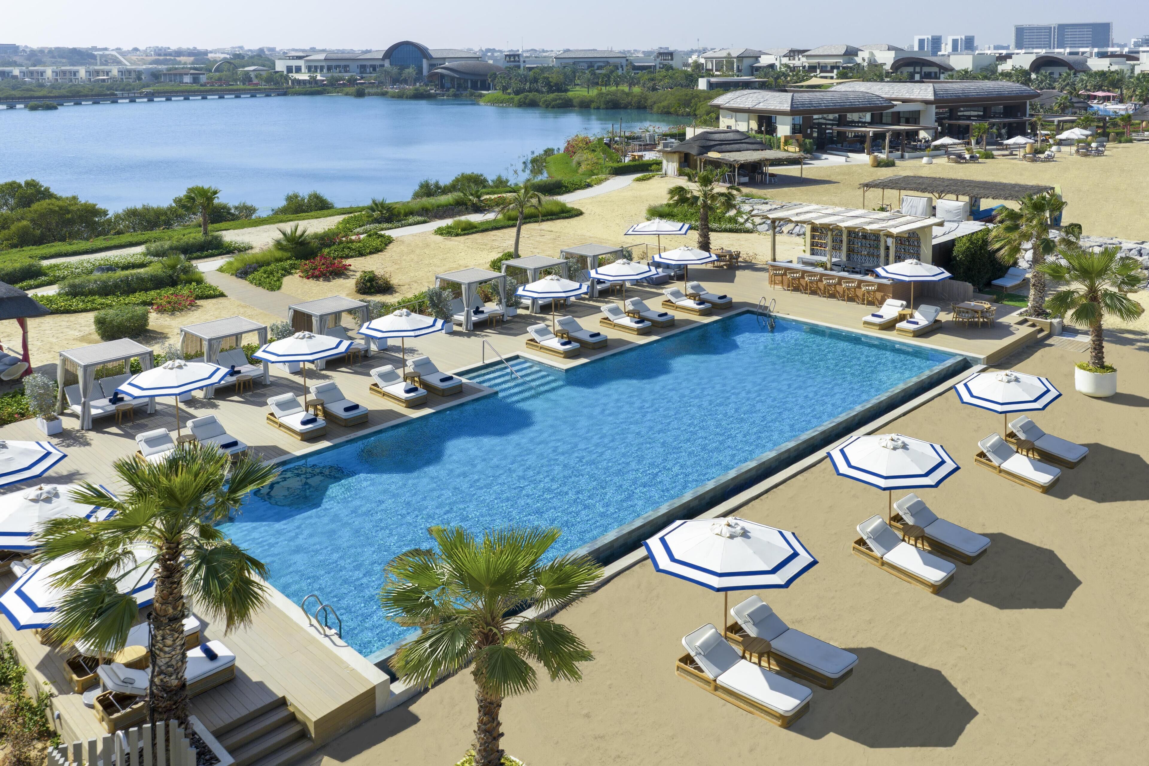 A large swimming pool surrounded by sun loungers and umbrellas, with a view of a beach and water in the background.