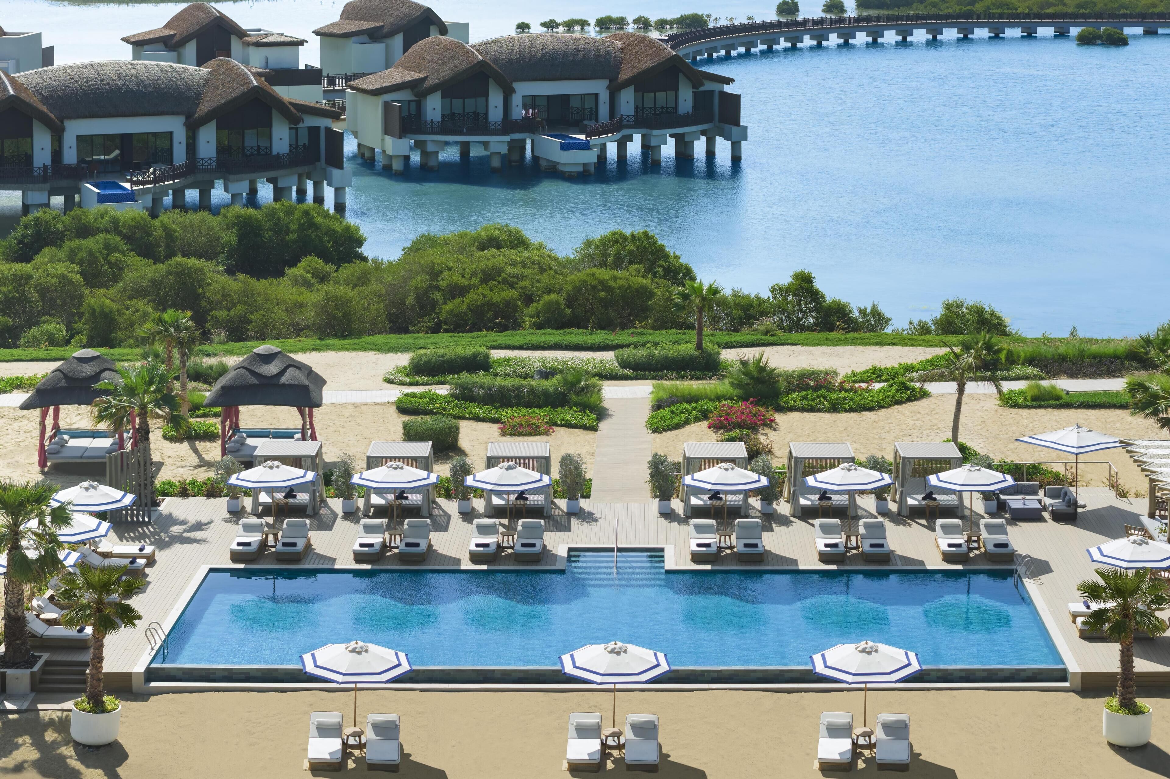 An infinity pool with lounge chairs and umbrellas, overlooking a body of water with overwater bungalows.