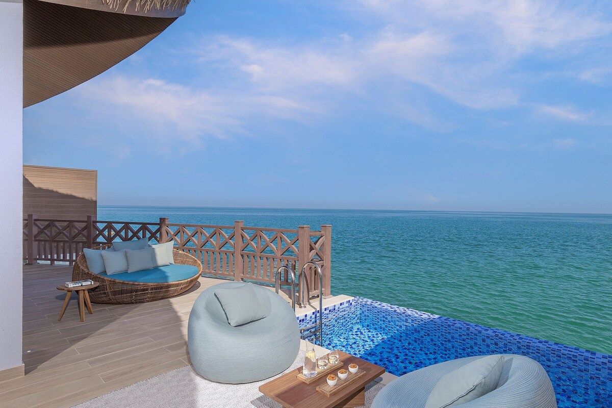 Un balcon avec un canapé en osier, un fauteuil poire et une petite table où sont posées des boissons, donnant sur une mer d’un bleu limpide.