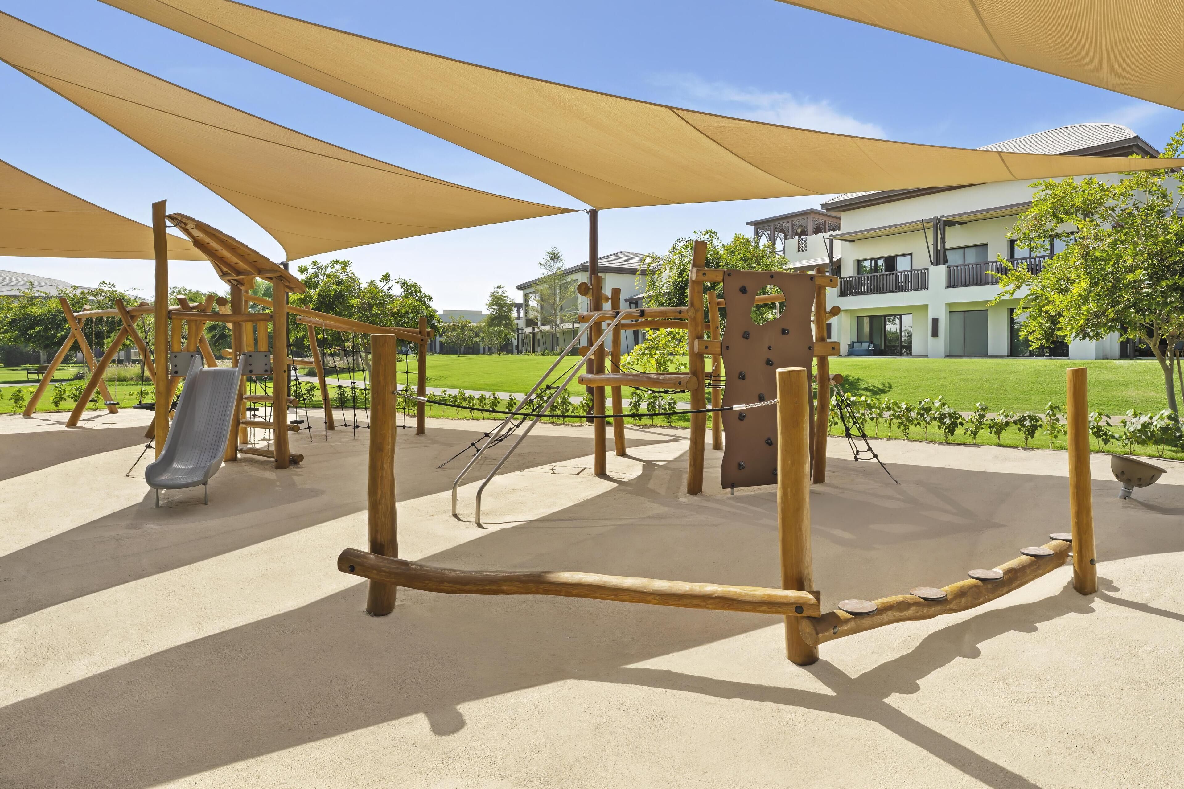 A playground with various wooden structures, including a slide and climbing frames, under large shade canopies.