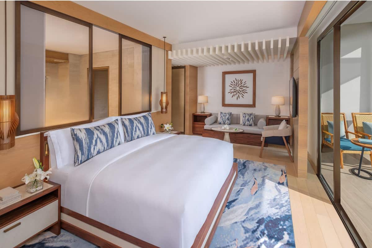 Una habitación de hotel moderna con una cama grande, una zona de estar con sofá y sillones, un ventanal amplio y un balcón con mobiliario exterior.