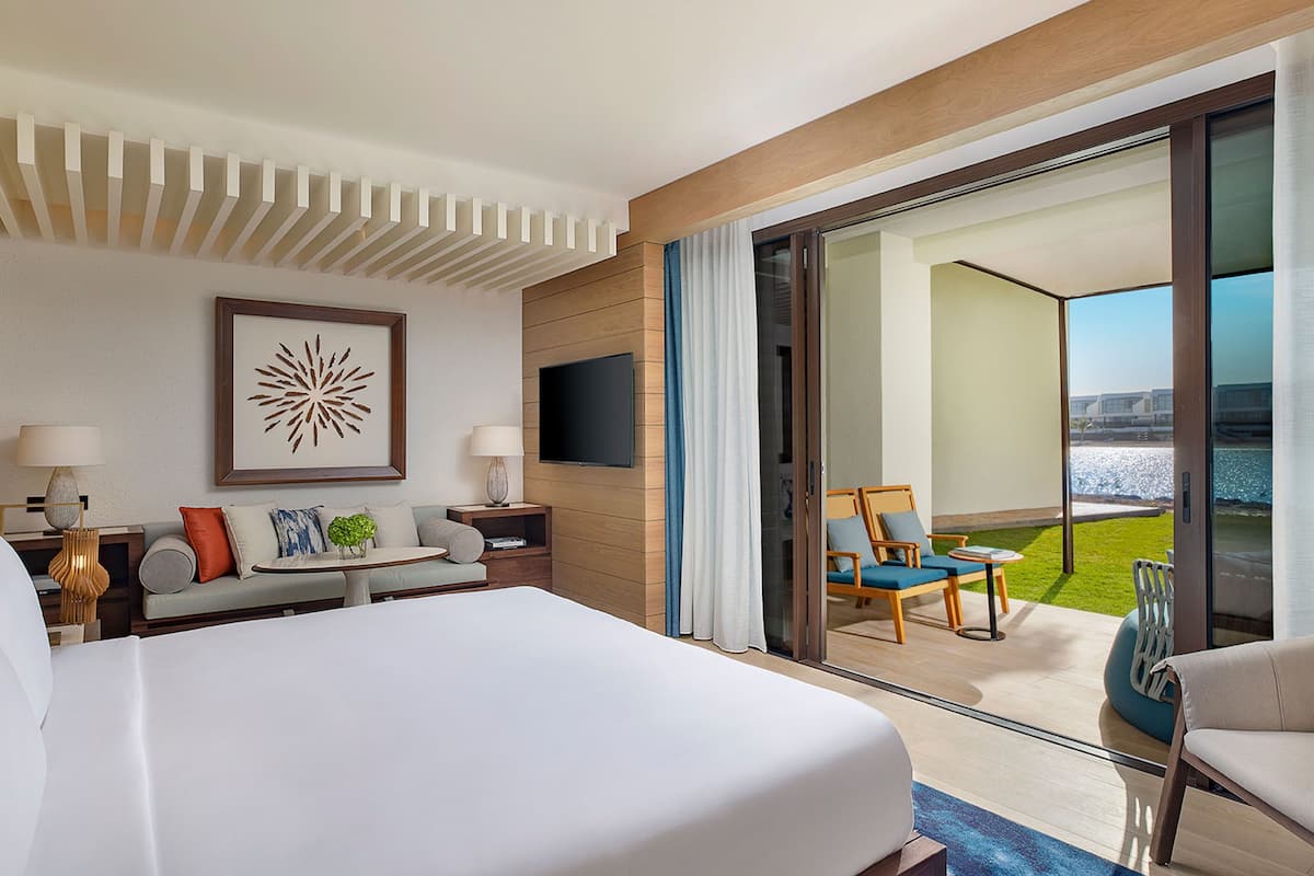 Habitación de hotel con una cama grande, un sofá, un televisor y un balcón con sillas y vista al mar.