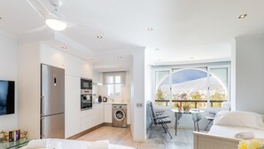 Una habitación de hotel moderna con cama, lavadora, zona de comedor con mesa y sillas, y balcón con vistas.