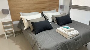 Cama bem arrumada com roupa de cama cinza e várias almofadas, mesa de cabeceira com abajur e janela que oferece iluminação natural.