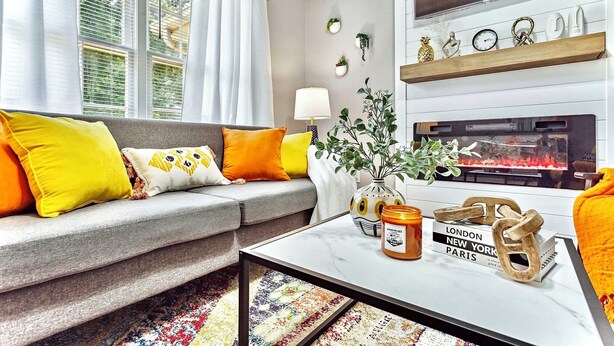 A living room with a grey sofa, yellow and white cushions, a white coffee table, a fireplace, and a TV mounted on the wall.