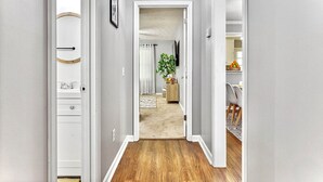 A hallway with a wooden floor, a white door on the left, and a dining area with chairs and a table visible in the background.