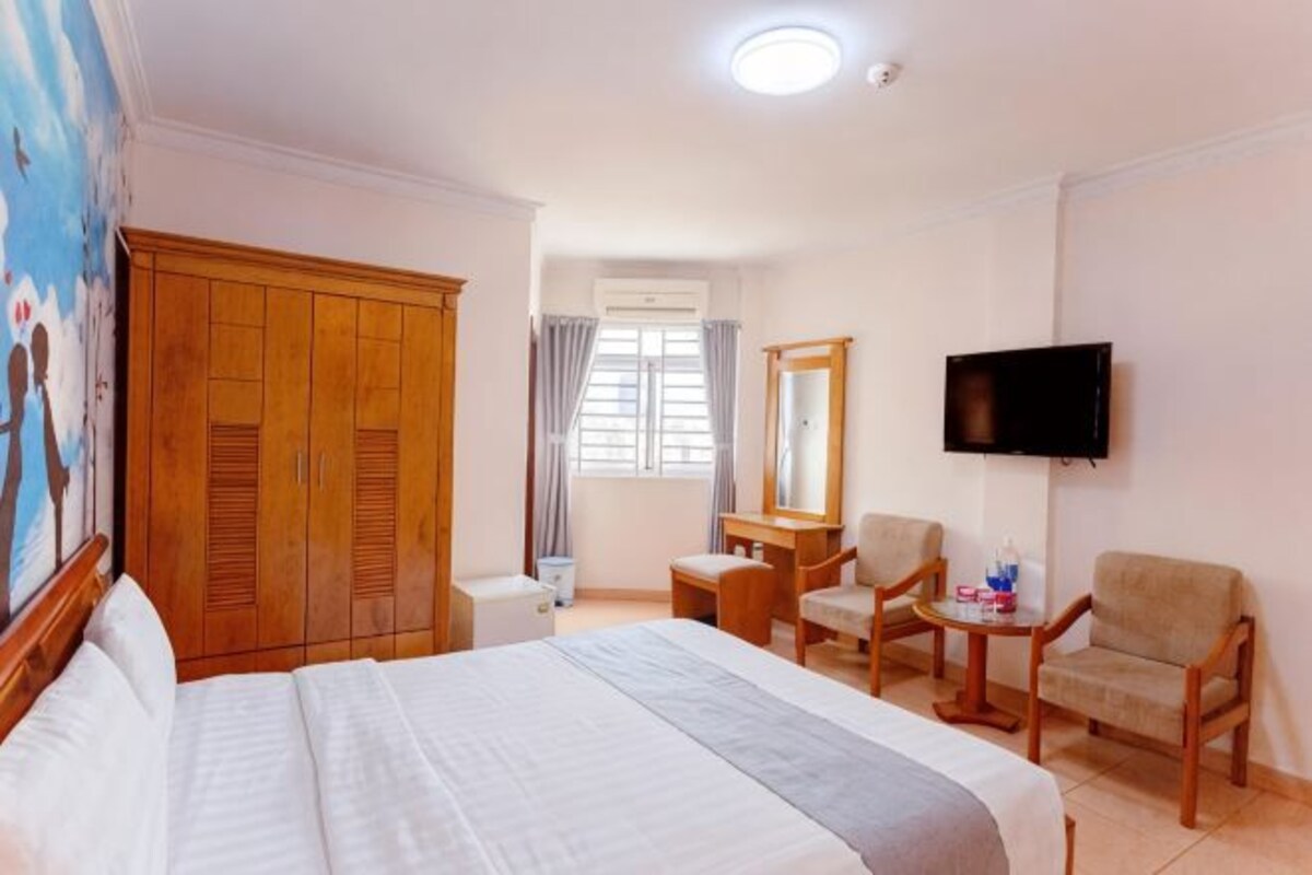 Ein Hotelzimmer mit Bett, Fernseher, Schreibtisch, zwei Stühlen, einem Kleiderschrank und einem Fenster mit Vorhängen.