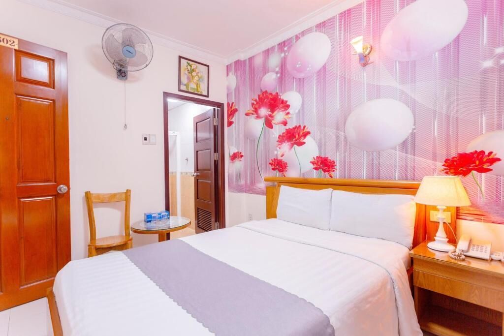 A hotel room with a bed, a bedside table with a lamp, a chair, a fan, and a door with a number.