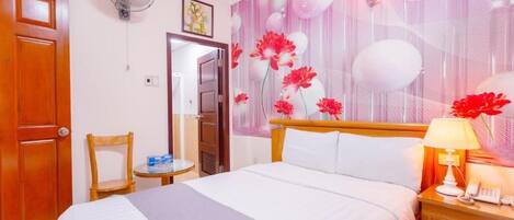 A hotel room with a bed, a bedside table with a lamp, a chair, a fan, and a door with a number.