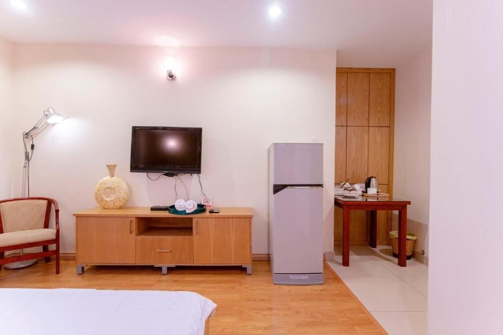 A hotel room with a bed, a TV mounted on the wall, a wooden cabinet, a refrigerator, a desk with a lamp, and a wooden wardrobe.