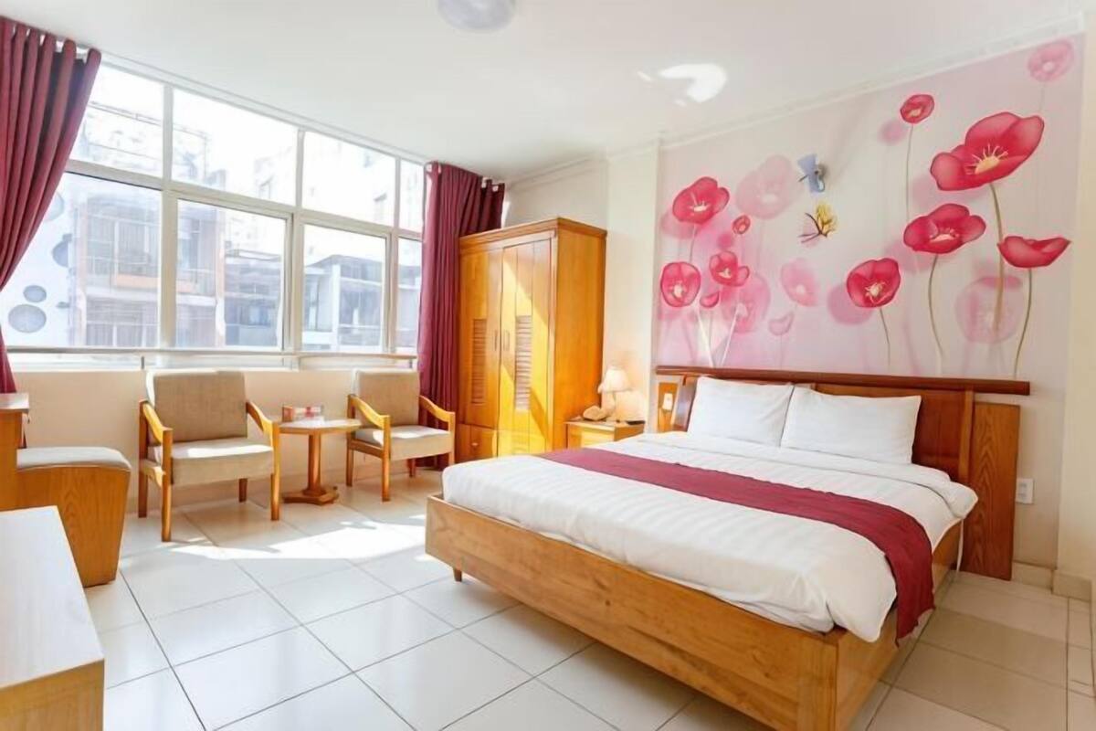 A hotel room with a large bed, a wooden wardrobe, a desk with a chair, a small table, and a window with curtains.