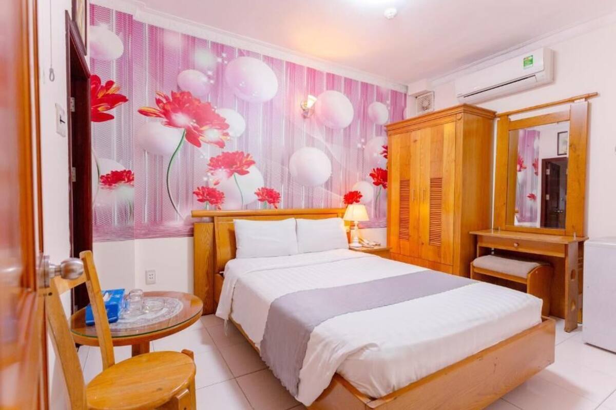 A hotel room with a bed, a wooden wardrobe, a small table with a chair, and a mirror.