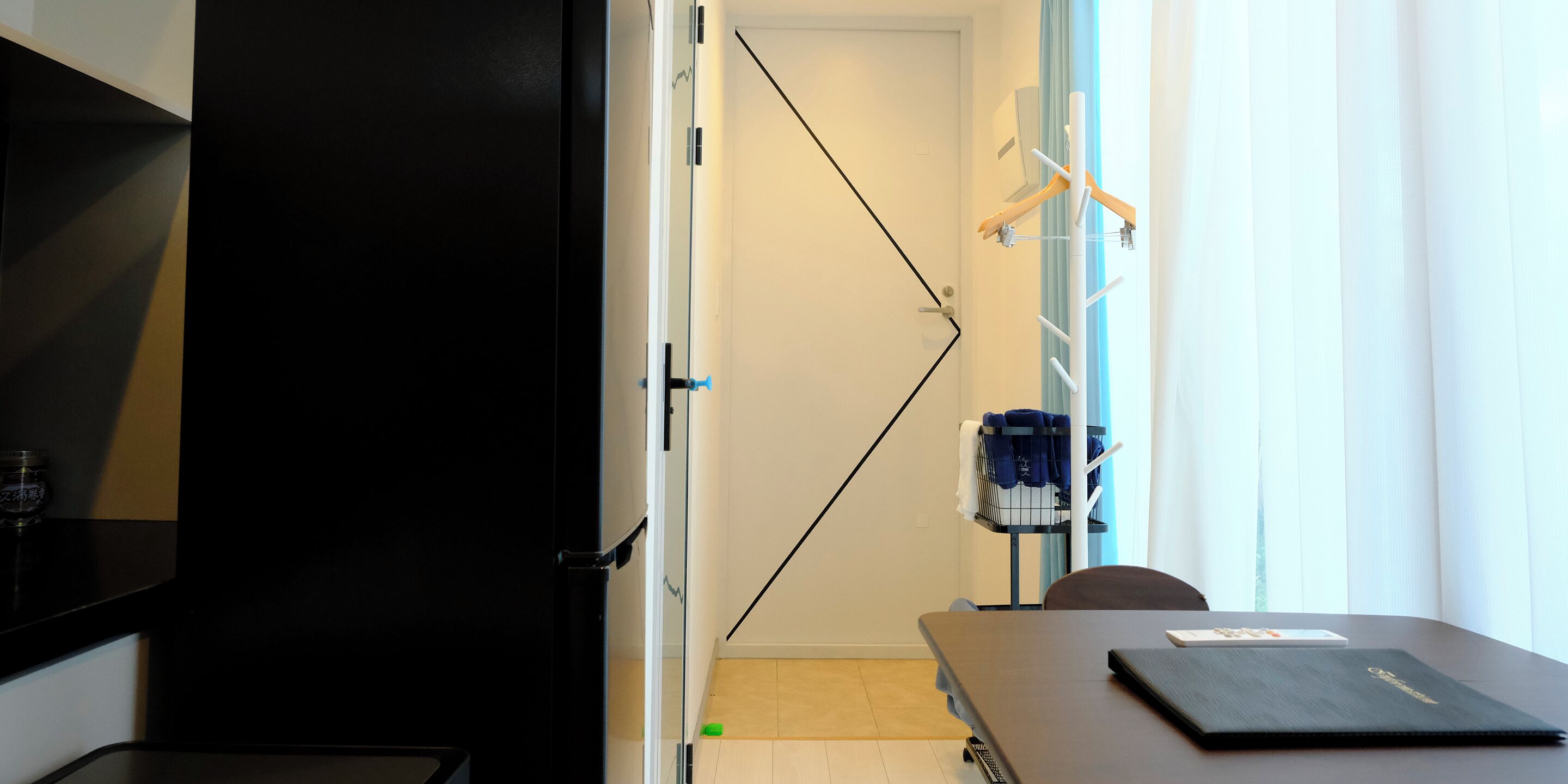 Una habitación con un escritorio negro, una silla, una mesita con una carpeta, una silla azul y un perchero con toallas.