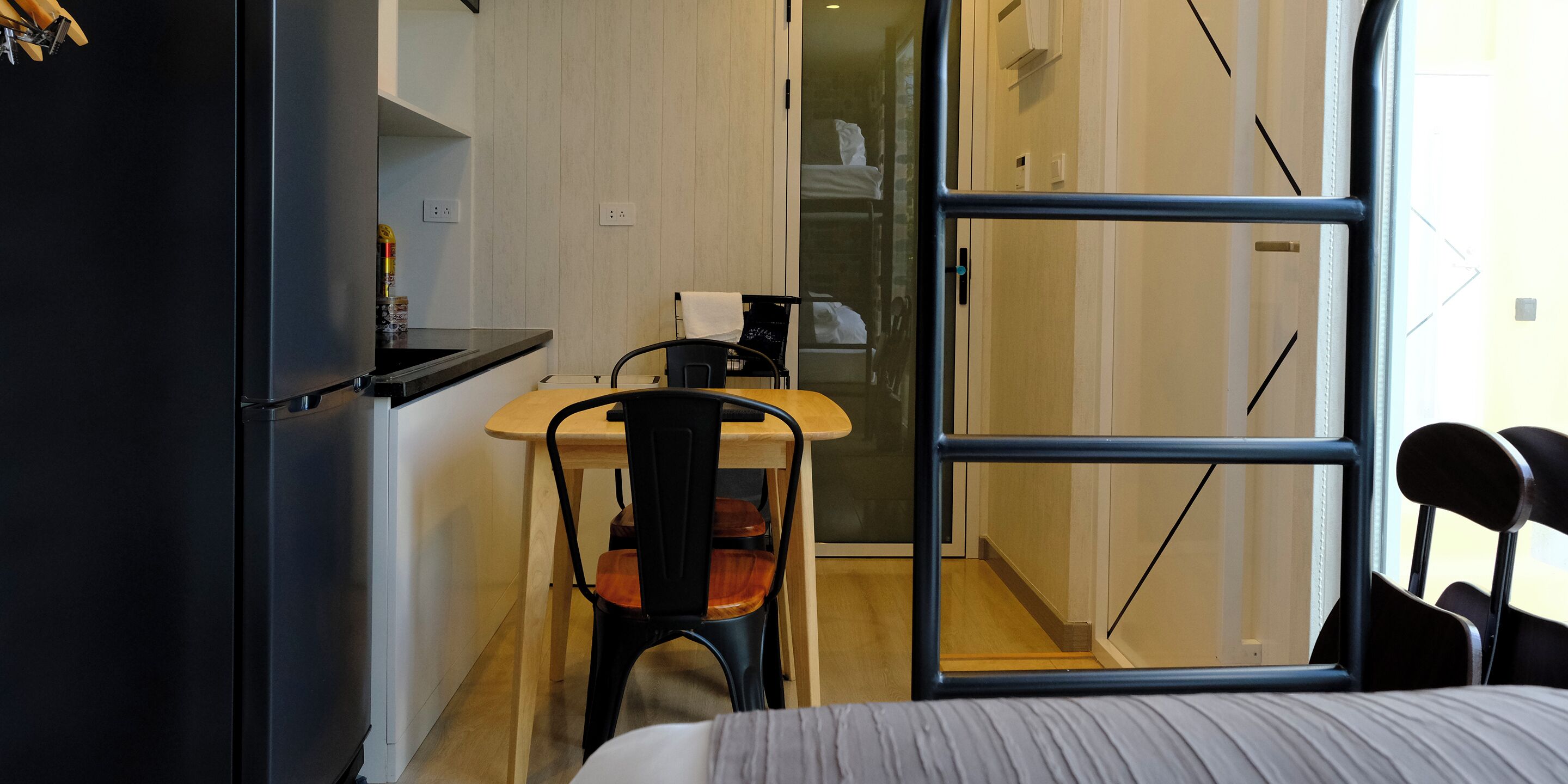 A compact room with a bed, a small table, and a chair. There is a window with a view of a staircase and a door leading to another room.