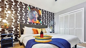 Un dormitorio con cama, cabecera, una obra de arte en la pared, una mesita de noche con una botella y un vaso, y una cesta sobre la cama.