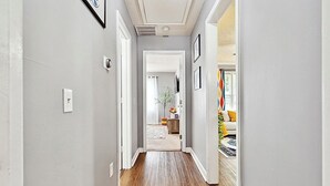 Un pasillo con piso de madera, paredes grises y detalles blancos. Hay cuadros enmarcados en las paredes y una puerta al final que da a una sala de estar con un sofá y un tapete colorido.