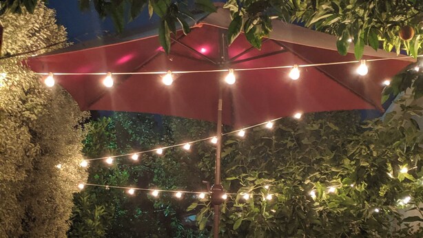 Eine Außenpatio mit Tisch und Stühlen unter einem roten Sonnenschirm, umgeben von Grünpflanzen und Lichterketten.