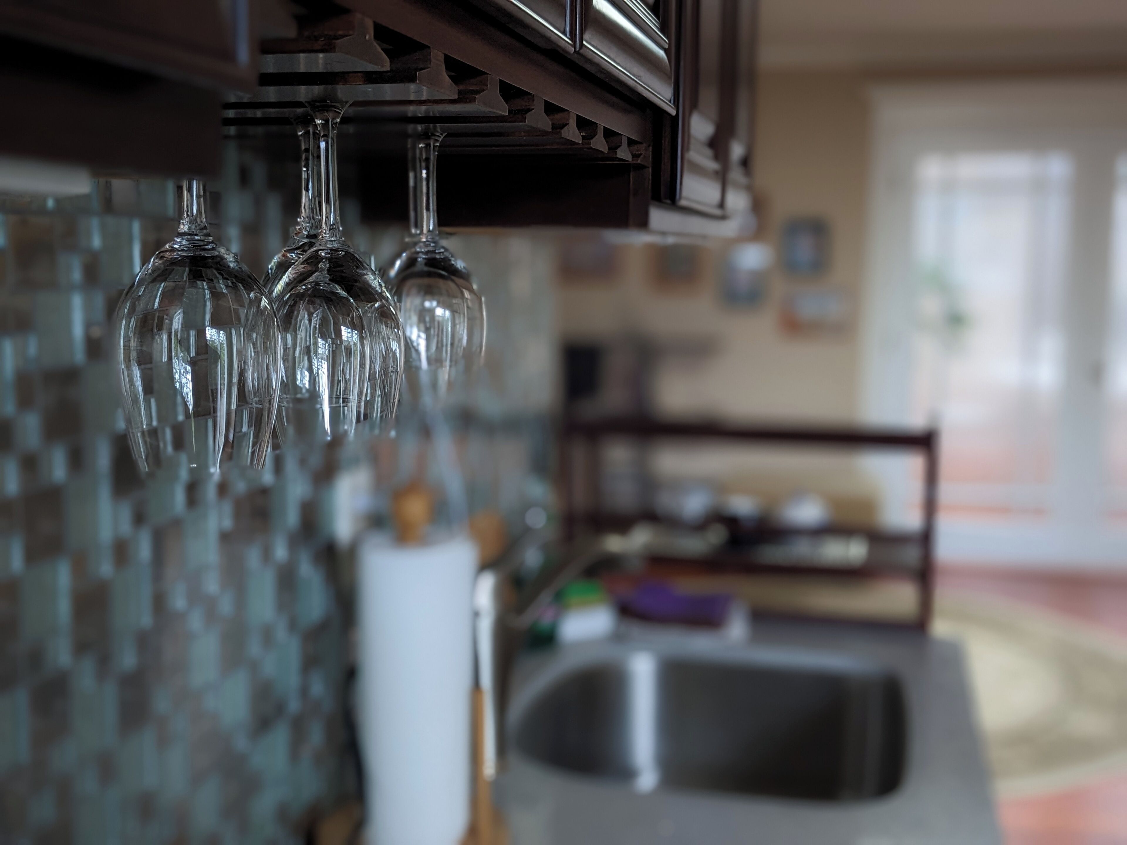 Una cocina con salpicadero de azulejos, un lavamanos de acero inoxidable y copas de vino colgando.