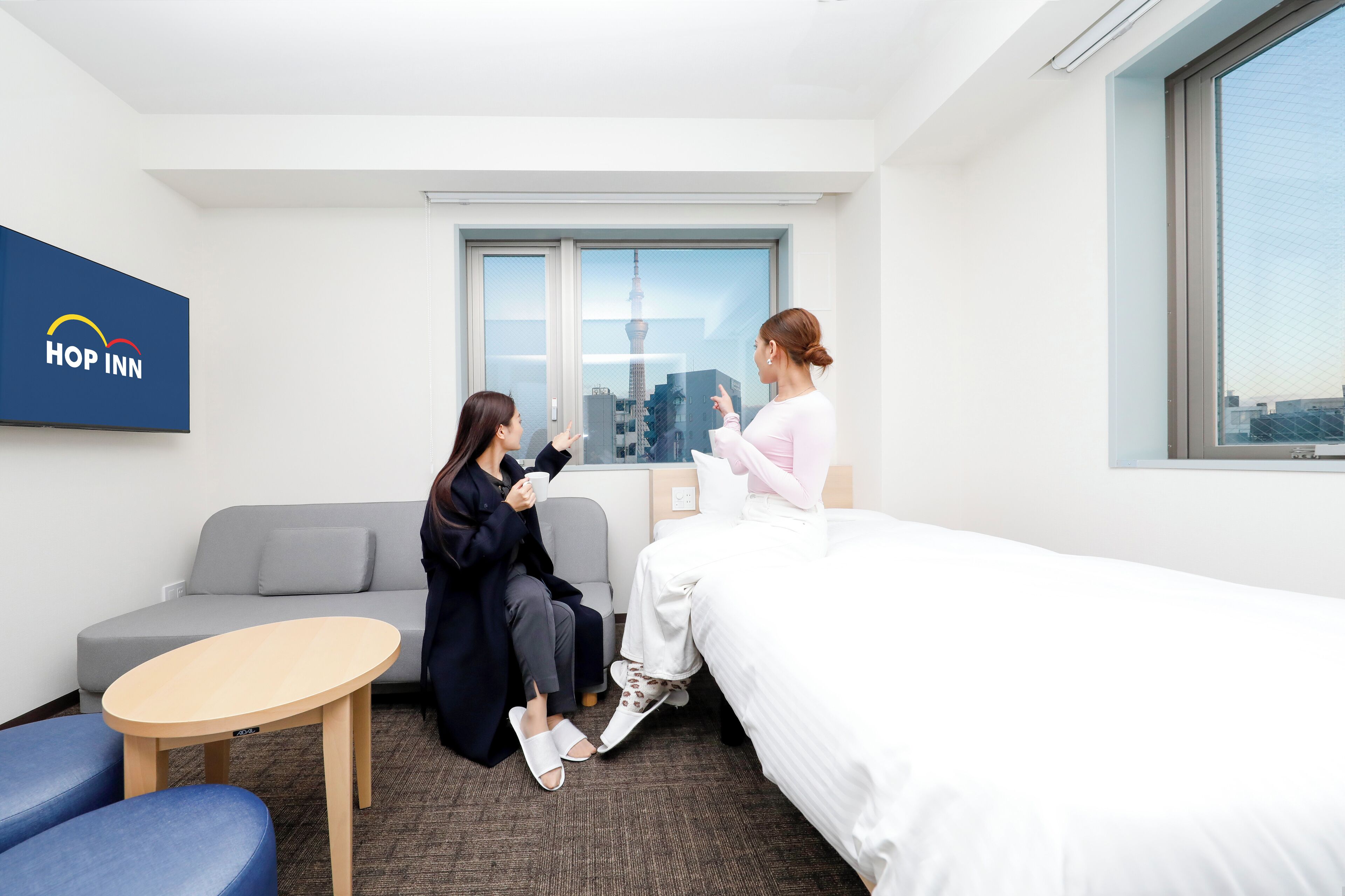 A modern hotel room with a bed, a sofa, a small table, and a window with a cityscape view.