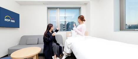 A modern hotel room with a bed, a sofa, a small table, and a window with a cityscape view.