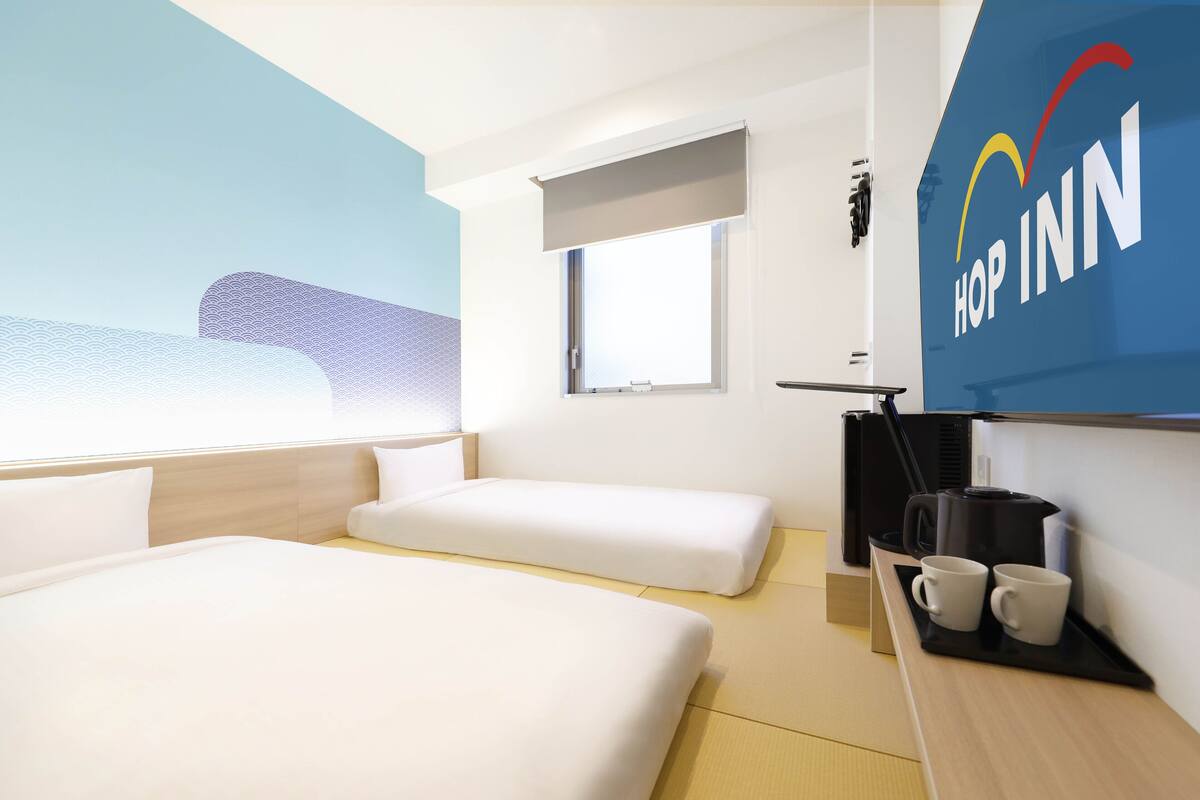 A hotel room with two beds, a window, a coffee maker, and a wall-mounted screen displaying the hotel name.