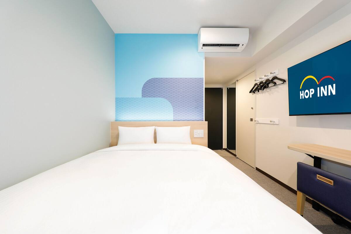 A modern hotel room with a large bed, a wall-mounted TV displaying the 'Hop Inn' logo, a wardrobe, and a door leading to another room.
