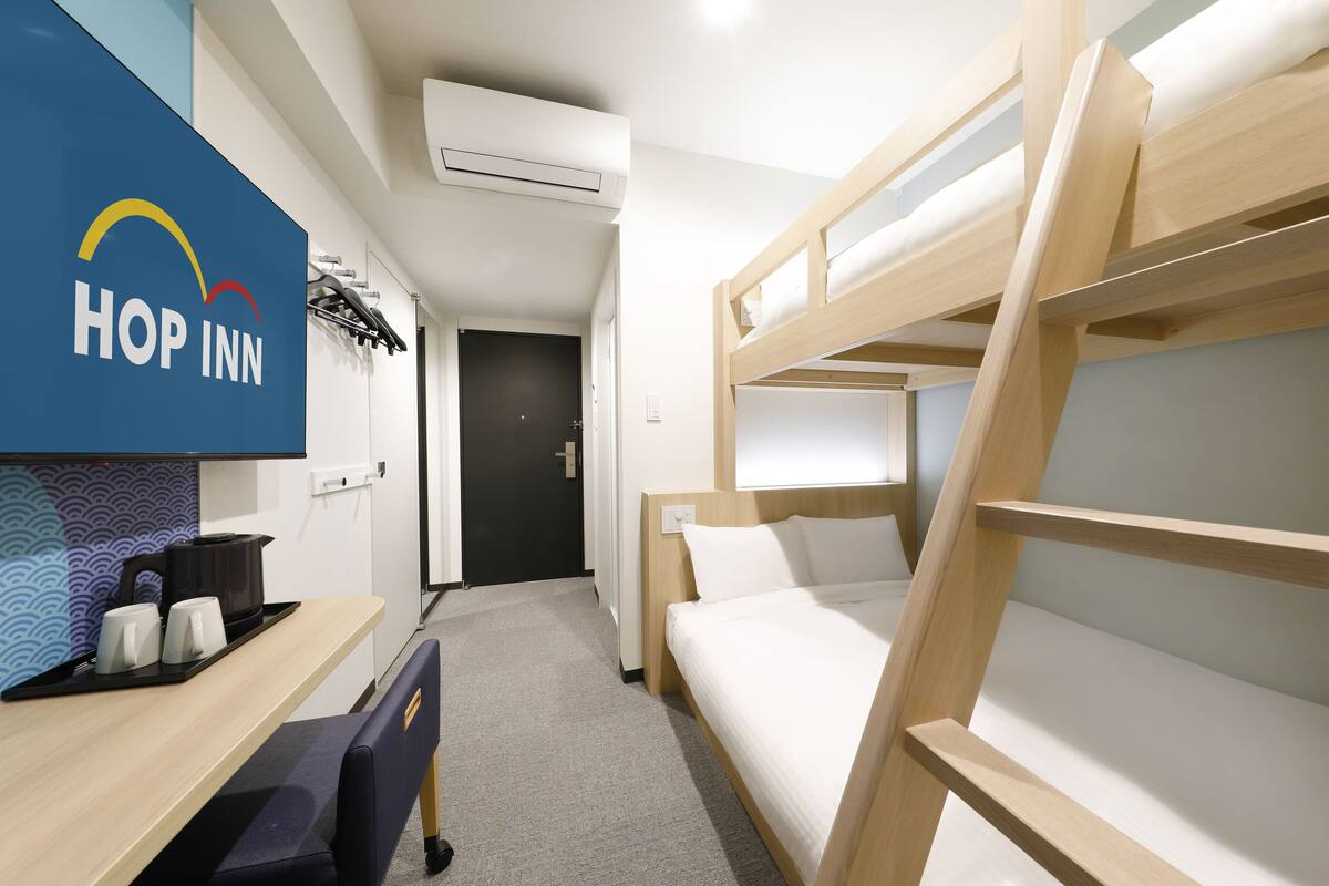 A bunk bed room with a desk and a chair, a wall-mounted air conditioner, and a wall with the "HOP INN" logo.