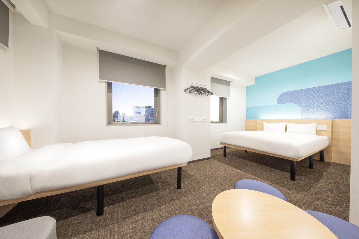 A hotel room with two single beds, a small table, and a window offering a city view.