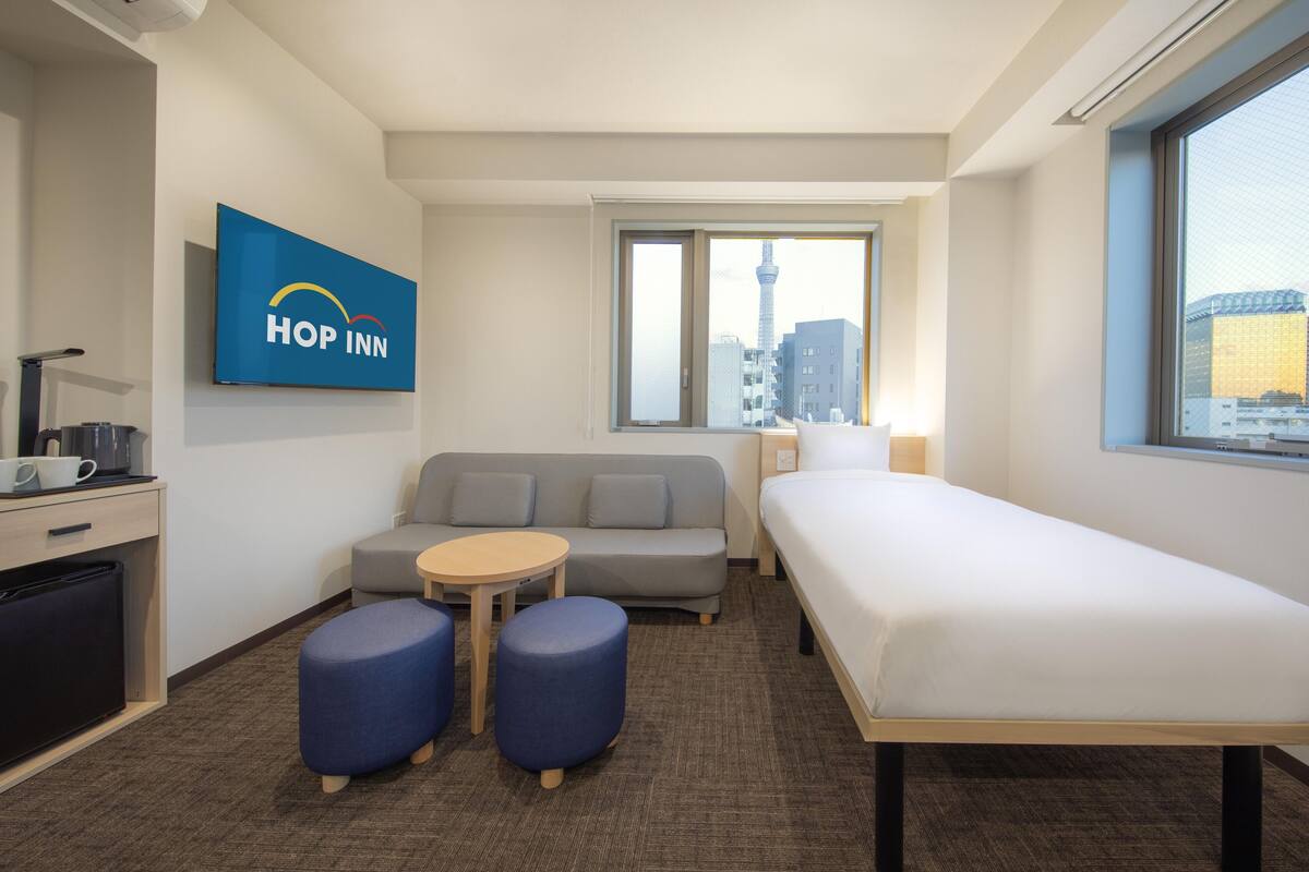 A compact hotel room with a sofa, a single bed, a small table, and a refrigerator. A large window offers a view of a cityscape, including a tower.